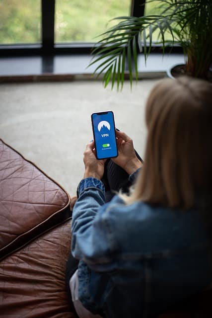 Woman sitting on a couch, camera is above her left shoulder as she sits facing away from the camera. She is holding up a phone that has a page loaded that says "VPN"