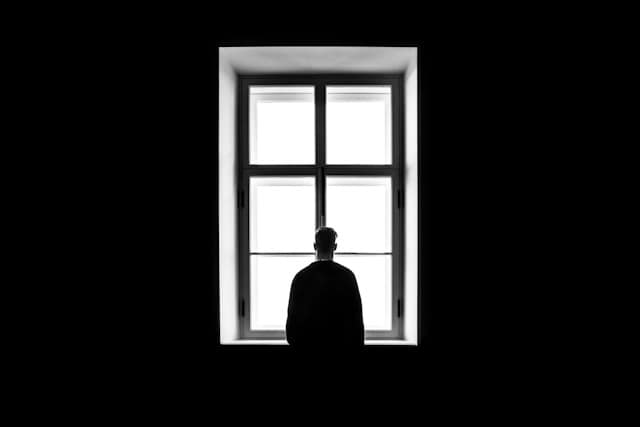 sasha-freemind-Pv5WeEyxMWU-unsplash man standing in front of a window in a dark-black room.