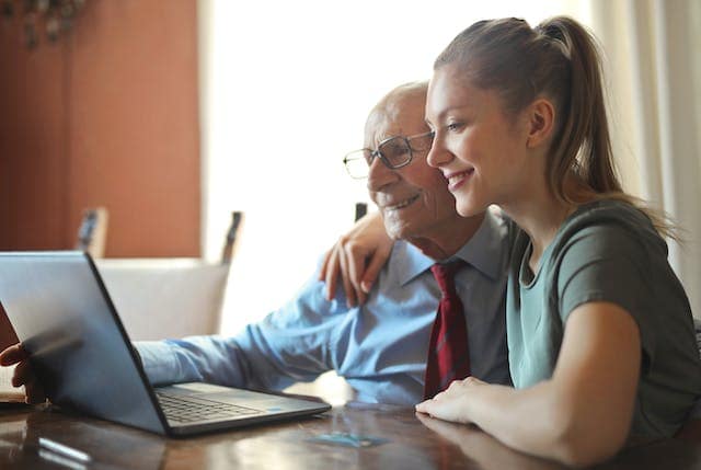 Young woman sitting next to a grandfatherly man, with her arm around his shoulder, both are smiling, they are sitting in front of a laptop both have smiles on their faces.