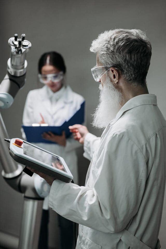 two scientists in a room with a robotic arm. The man in the foreground is holding a tablet-like screen in his left hand while the other is outstretched about towards the robot. The woman is blurred in the background and his writing in a binder.