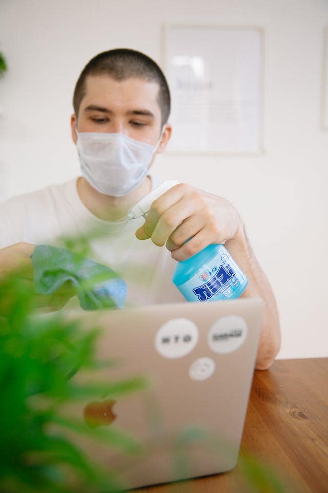 Man sitting at a table with a laptop with a plant in the blurry foreground. He is holding a rag and a bottle of cleaner which he is spraying on the rag.