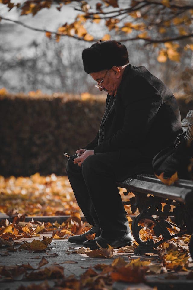 Man in a coat, scarf, and a Russian fur hat sitting on a park bench in fall, he's looking down at the phone in his hands between his legs.