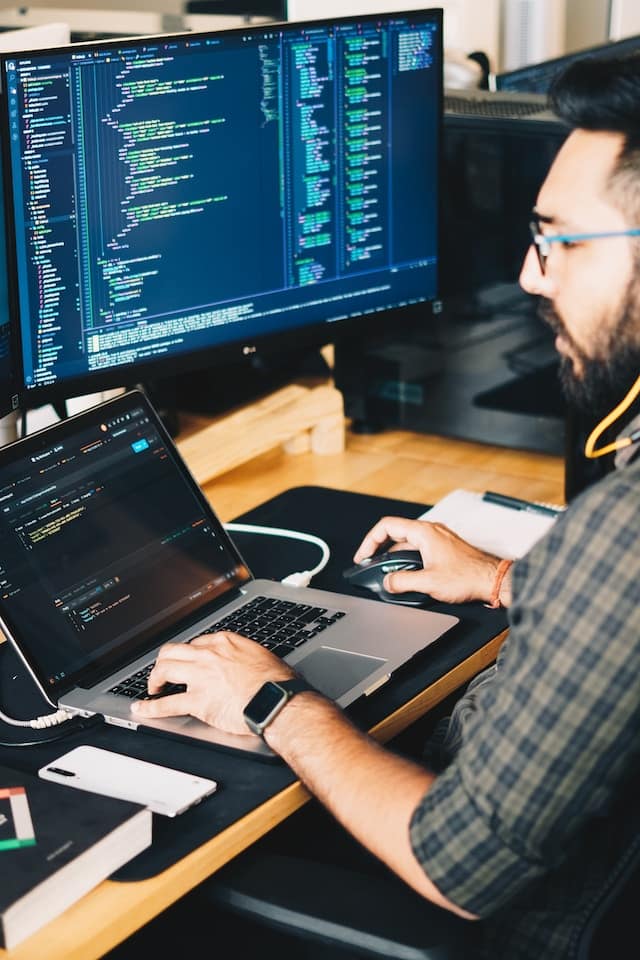 Man sitting at a desk with a laptop and another monitor, on the screens are code.