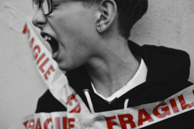 Young boy up against a wall, his face is pointing to the left and it looks like he's crying out in frustration, eyes are shut tight and his mouth is open. There's tape pinning him to the wall, the tape reads 