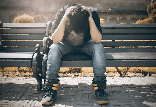 Man sitting on a park bench, hunched over looking down between his legs with his elbows on his knees and his hands on the back of his head.