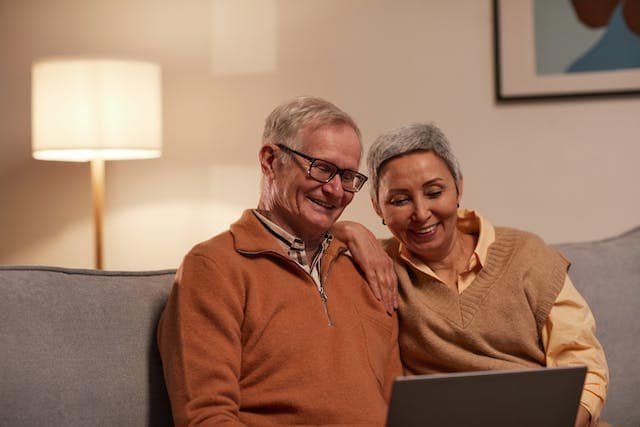 pexels-marcus-aurelius-6787970 Two seasoned adults sitting on a couch, they are both smiling and looking down at the screen of a laptop.