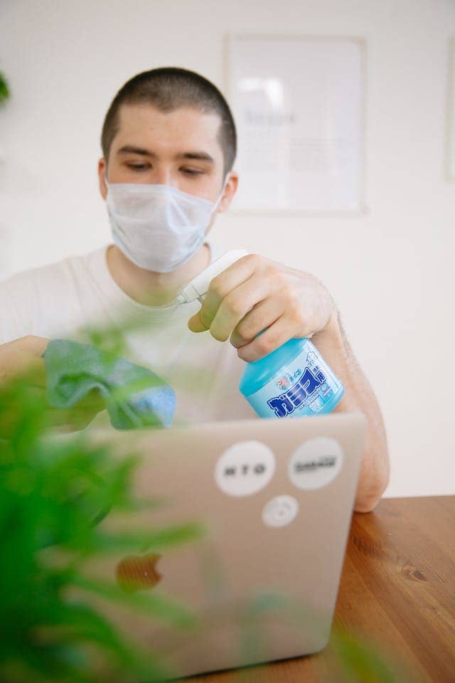 Man sitting at a table with a laptop with a plant in the blurry foreground. He is holding a rag and a bottle of cleaner which he is spraying on the rag.