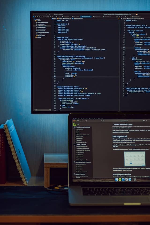 On monitor sitting on a stand with a laptop in front so they person using the laptop can also see the monitor behind. Both Screens have code on them.